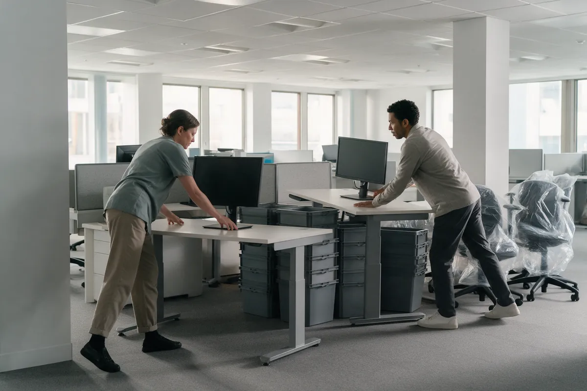 Two people moving desks and computer monitors across a bright open-plan office