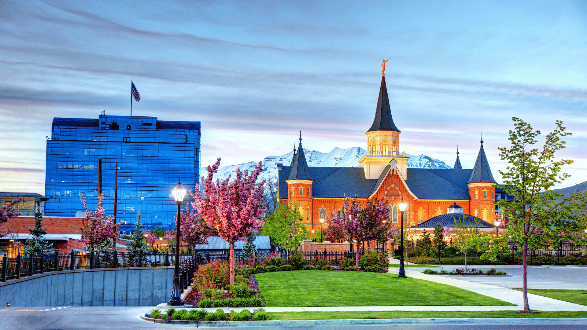 The Provo City Center Temple and downtown Provo at dusk, with spring trees in bloom and snow on the mountains behind
